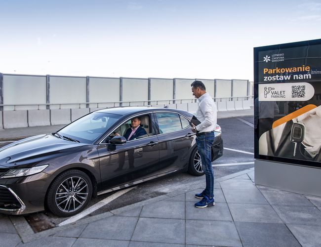 Parkservice – Chopin-Flughafen. Der Fahrer in einem dunklen Auto spricht mit einem Mann. Nebenan steht ein großes Informationsschild.