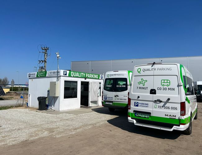 Q-Parking – Kontener biurowy i dwa białe busy. Teren jest żwirowy i nieogrodzony.