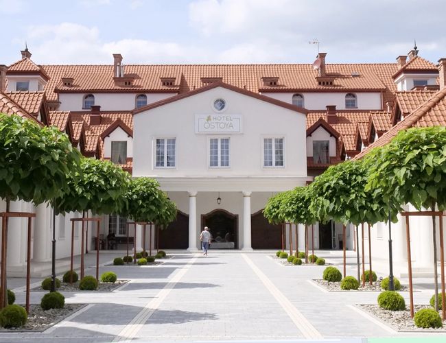 Parking Zielony Kompleks Dwór Ostoya – Budynek hotelu z czerwonym dachem. Wzdłuż drogi rosną symetryczne drzewa. Na końcu dziedzińca jest wejście.