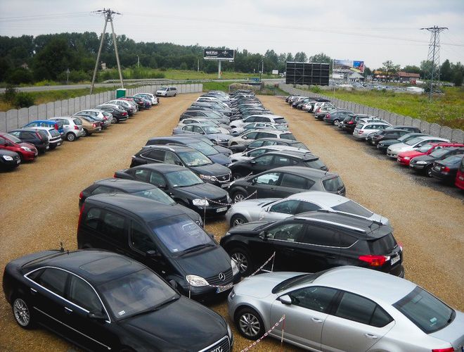 TRAVEL Parking - Wiele aut stoi na żwirowym placu. Parking ogrodzony jest siatką. W tle widać roślinność.