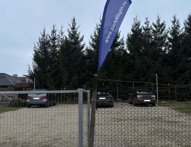 Ogrodzony parking Park4flight Maramureș z widoczną niebieską flagą reklamową przy bramie wjazdowej oraz kilkoma zaparkowanymi samochodami na żwirowym terenie, w otoczeniu wysokich drzew.