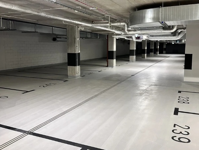 Garaje de Estacionamiento Supremo – El estacionamiento subterráneo está vacío y bien iluminado. Hay números de espacios en el suelo. Las tuberías son visibles en el techo.