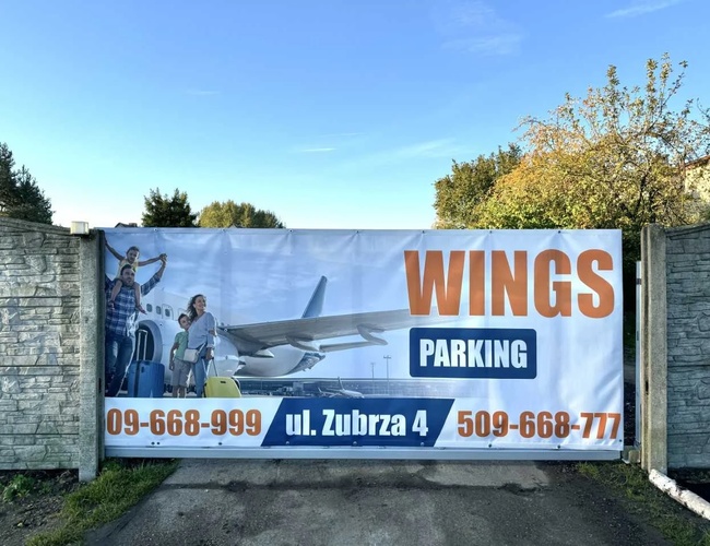 Parking Wings - Banner publicitario en una valla de concreto. La gráfica muestra una familia con equipaje junto a un avión. Árboles visibles al fondo.