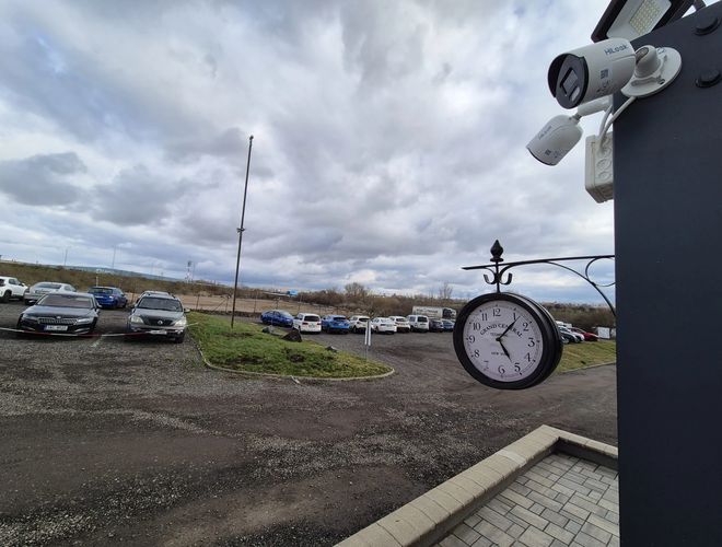 HostParking - Veľké parkovisko s niekoľkými autami. Viditeľné hodiny a kamery. Zamračená obloha na pozadí.