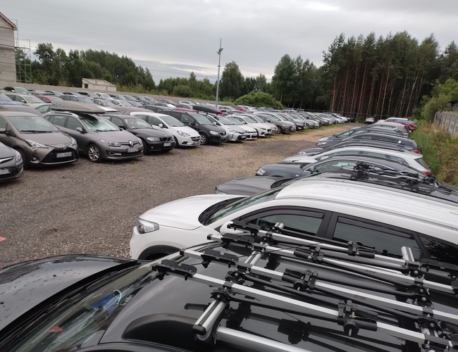Gran Estacionamiento – muchos coches personales estacionados. El estacionamiento está en una superficie de grava. Está rodeado de árboles y una cerca.