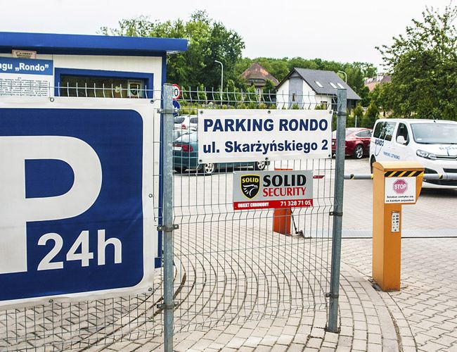Parking Rondo – ogrodzony teren z monitoringiem. Wjazd przez szlaban. Znak informuje o całodobowej ochronie.