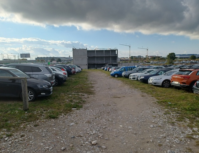 Parking Goliat – Szutrowa nawierzchnia dzieli rzędy zaparkowanych aut. Obok placu widoczny jest piętrowy budynek. Na horyzoncie widać dźwigi.