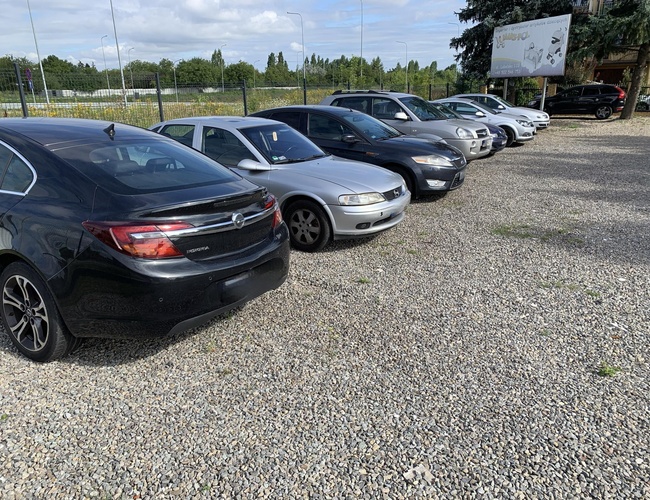 Parkovanie Max A1 - Rada áut stojí na štrkovom dvore. Parkovisko je ohradené sieťou. V pozadí je vidieť billboard.