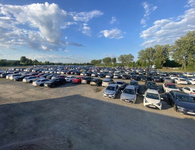 Smart Premium Parking - Obszerny parking pełen samochodów. Nie ma zadaszenia. Otoczony jest drzewami pod niebieskim niebem.