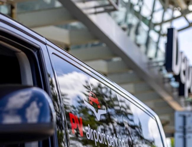 Samochód transferowy PV Airport Parking zaparkowany przy terminalu lotniska, z widocznym oznaczeniem firmy na pojeździe.