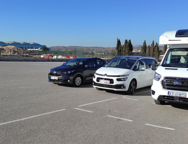 Parking Odkryj-Auto - Na parkingu stoją trzy pojazdy. W tle widać wzgórza. Teren jest ogrodzony siatką.