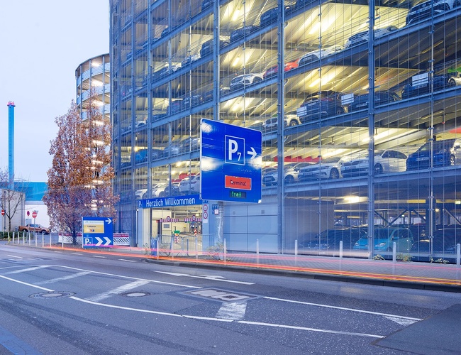 Wielopoziomowy parking P7 widziany od strony ulicy z widoczną drogą dojazdową oraz oznakowaniem parkingowym.