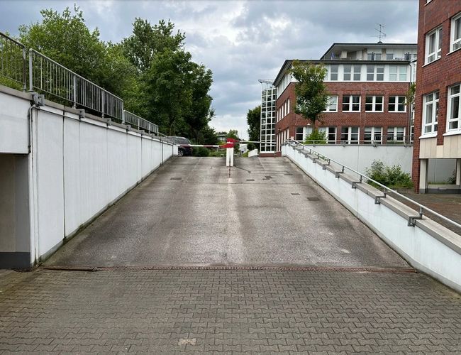 Parking Marspark Outdoor Valet – Wjazd podjazdem ograniczonym barierką. Widoczne drzewo i budynek z czerwoną cegłą. Niebo jest pochmurne.