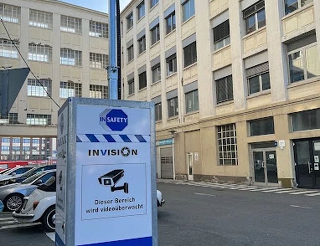 Parking Parkfinder24 Valet – Budynek stoi przy placu parkingowym. Na pierwszym planie jest szary kontener z kamerą. Obok widać zaparkowane samochody.