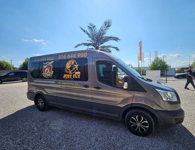 Just Fly Parking - Widoczny jest srebrny van z logo firmy. Samochód stoi na żwirowym placu pod palmą. W tle widać drzewa i przechodnia.