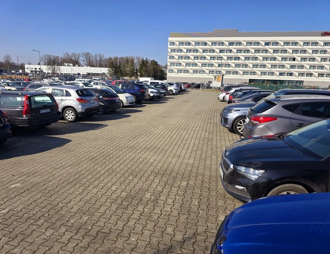 Parkovisko Olo Car Park – mnoho áut stojí na nádvorí. Pozemok je dláždený kockou. V pozadí je vidieť budovu.