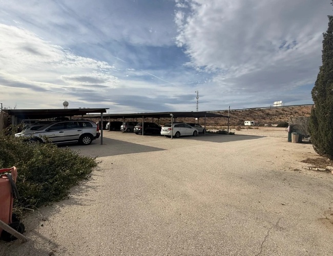 Szeroki parking z kilkoma zaparkowanymi autami, w tle niskie zabudowania i pagórkowaty teren pod błękitnym niebem.