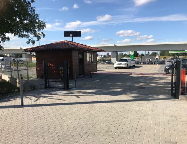 Autofahrzeug Flughafen Selbstbedienung – Der Parkplatz ist eingezäunt und befestigt. Der Zugang wird durch eine Schranke neben dem Holzwagen geschützt. Auf dem Platz stehen Autos.