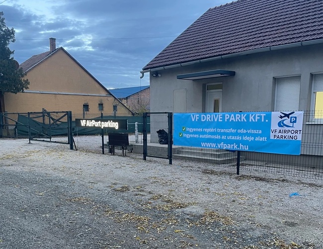 Widok na ogrodzony teren z budynkiem i banerem firmy przy wejściu, pochmurne niebo w tle.