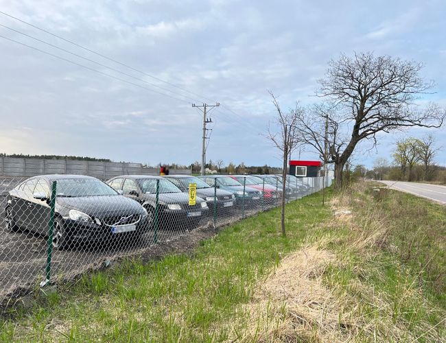 TRIP Parking - Estacionamiento con cercado de malla y coches. El terreno está adyacente a una carretera asfaltada. Al lado hay árboles.