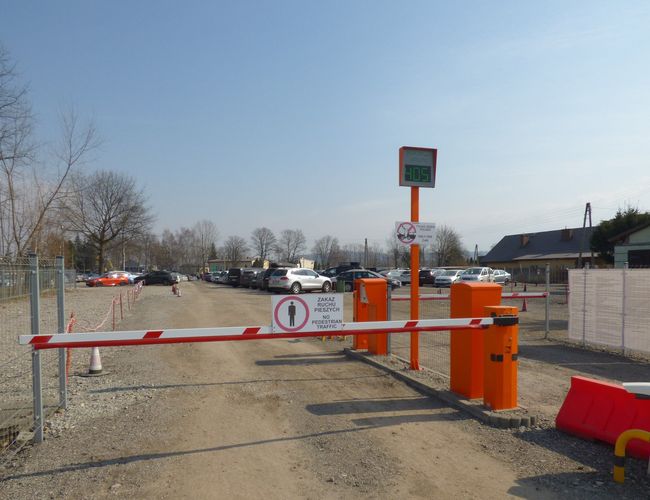 Krk Parking Lotnisko – Szlaban przy wjeździe i widoczne auta. Utwardzona droga prowadzi przez parking. Teren jest ogrodzony.