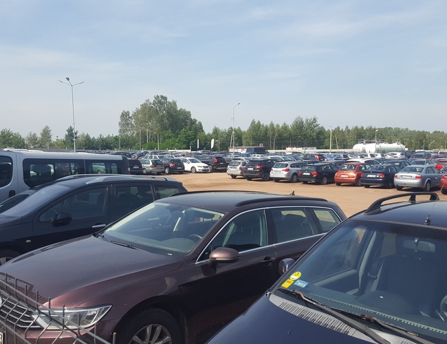 AAP-66 – na parkingu stoi wiele samochodów. Teren jest ogrodzony i przestronny. W tle rosną drzewa.