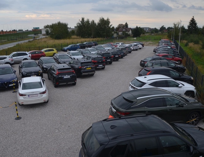 Royal Parking - Plocha je štrková a ohradená. Veľa zaparkovaných áut. Na pozadí cesta a stromy.