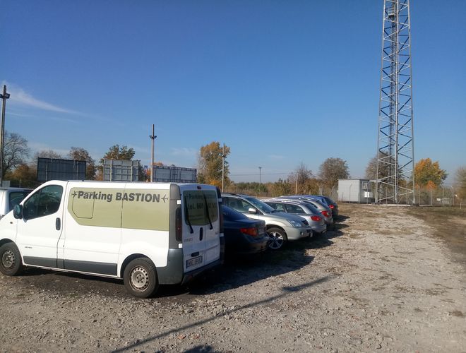Parkoló Bastion - A parkoló autók sora a fehér busz mellett áll. A terület nem aszfaltozott, és magas kerítéssel van körülvéve.