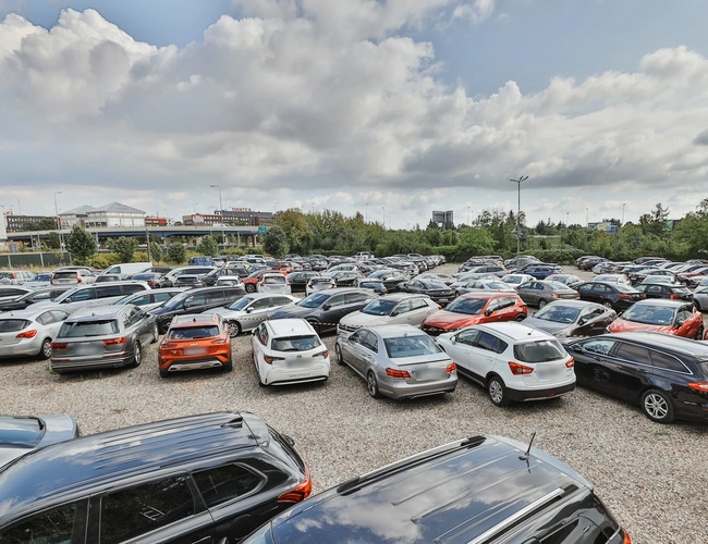 Smart Supreme Parkovanie - Veľké, zaplnené parkovisko s viacerými autami. Areál je otvorený a neobkľúčený. Na oblohe sú viditeľné oblaky.
