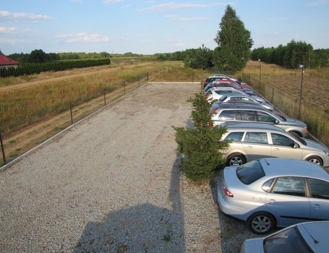 Parking Auto Park – Plac żwirowy z szeregiem zaparkowanych samochodów. Teren jest ogrodzony z widocznymi lampami.