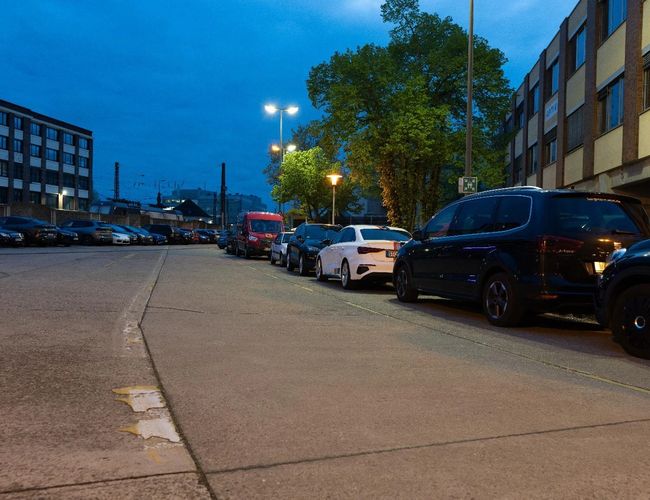 Parking Clean Valet Parkservice – na parkingu stoją zaparkowane samochody. Teren jest oświetlony latarniami. W tle widać budynek i drzewa.