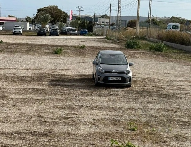 Samochód zaparkowany na nieutwardzonym parkingu, w tle inne auta, słupy energetyczne i roślinność.