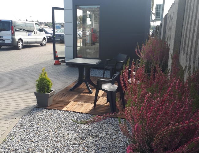 WIZ-PARK - Mesa con sillas en una plataforma de madera. Al lado, plantas en macetas junto al camino de grava. Al fondo, un edificio.