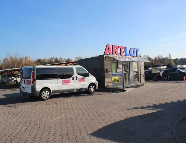 ArtLot 2 – el autobús está frente al edificio con un gran letrero. El aparcamiento es espacioso y está bien iluminado. Al fondo se pueden ver coches estacionados.