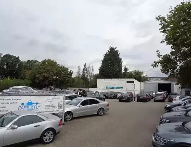 Parking Park&Fly Düsseldorf - Teren jest pełen zaparkowanych samochodów. Otoczony drzewami, w tle widać budynek.