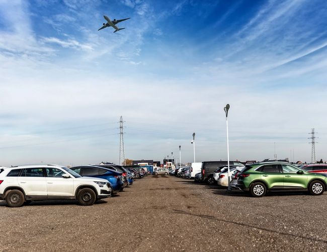 Parking Okęcie Południe - Samochody stoją na żwirowym placu. Lampa oświetleniowa przy drodze. Na niebie leci samolot.