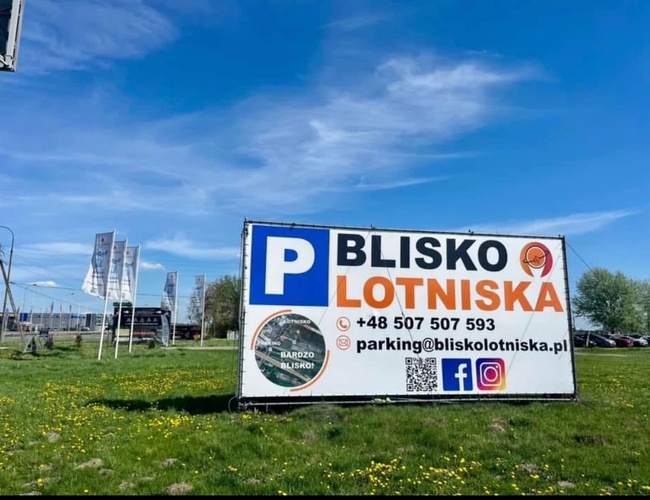 Parking Blisko Lotniska – Rząd samochodów stoi na żwirowym placu. Teren jest otwarty, z widocznymi flagami reklamowymi. W tle widać drzewa.