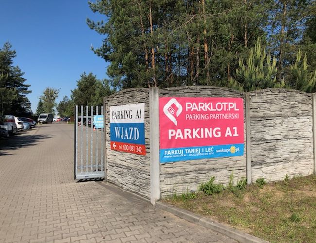 Parking A1 - betonowe ogrodzenie i otwarta brama. W tle widać samochody. Parking jest otoczony drzewami.