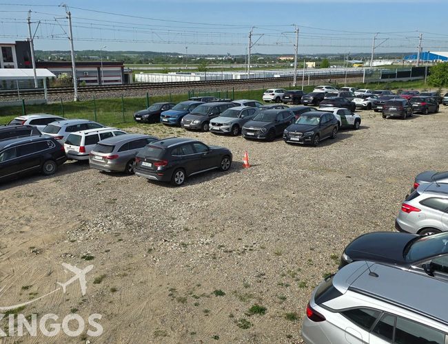Parking Parkingos - Żwirowa nawierzchnia z zaparkowanymi samochodami. W tle są tory kolejowe i budynki. Parking jest ogrodzony.