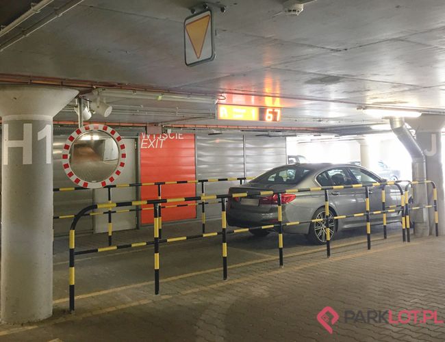 Oficjalny Parking Lotniska P2 – Auto stoi w podziemnym parkingu. Są barierki ochronne i znaki. Widać wyjście ewakuacyjne.