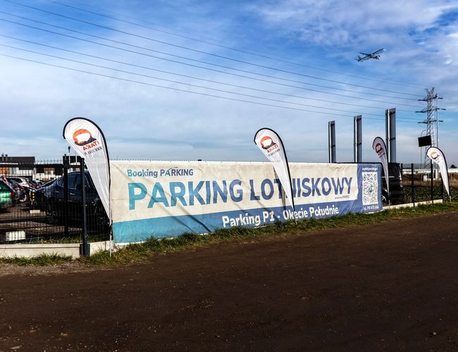 Parking Okęcie Południe - Ogrodzony teren z wieloma zaparkowanymi samochodami. Na płocie znajduje się duży baner informacyjny.