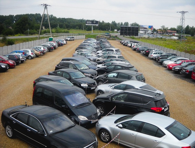 TRAVEL Parking - Muchos coches están en el área de grava. El estacionamiento está cercado con una malla. Al fondo se puede ver vegetación.