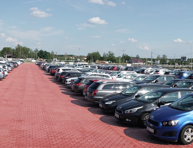 MEGA Parking - En la plaza roja hay muchos coches. El área está rodeada de árboles y farolas.
