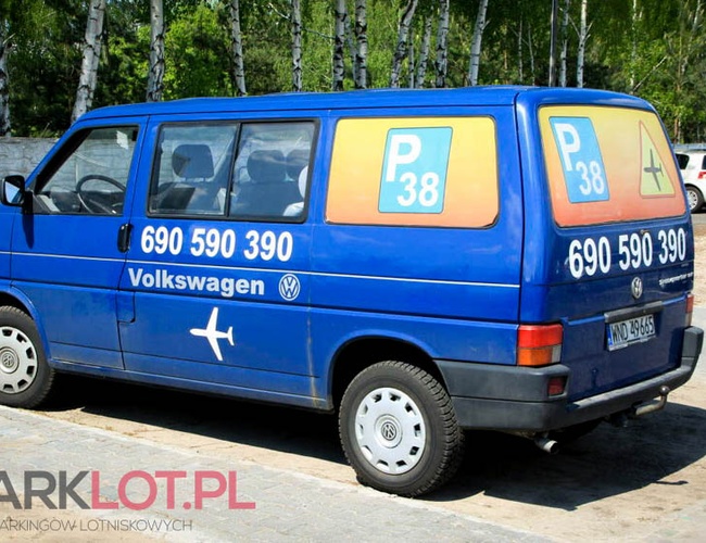 Parking P38 - Modrý autobus s telefónnym číslom stojí na námestí. V pozadí sú viditeľné stromy a zaparkované autá.