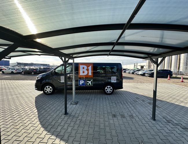 B1 Parking zadaszony - Pod wiatą stoi oznakowany pojazd. W tle widać zaparkowane samochody. Nawierzchnia jest wyłożona kostką brukową.