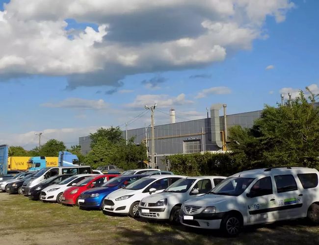 Rząd kolorowych samochodów zaparkowanych przy trawniku obok budynku przemysłowego w słoneczny dzień.