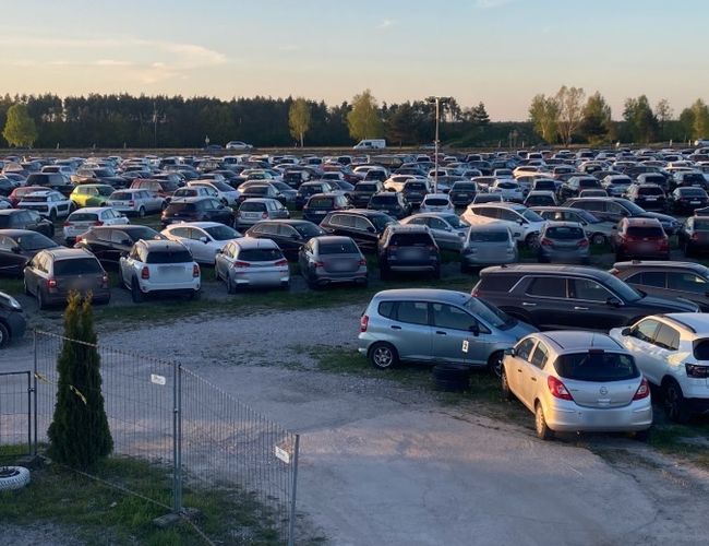 Aparcamiento bajo los molinos de viento – muchos coches están estacionados en un gran aparcamiento. El entorno es verde, con filas de árboles al fondo.