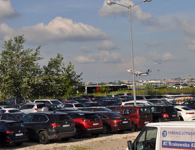 Parking Lotnisko – Zaparkowane samochody widoczne na dużym placu. W tle startuje samolot. Teren jest ogrodzony i oświetlony.