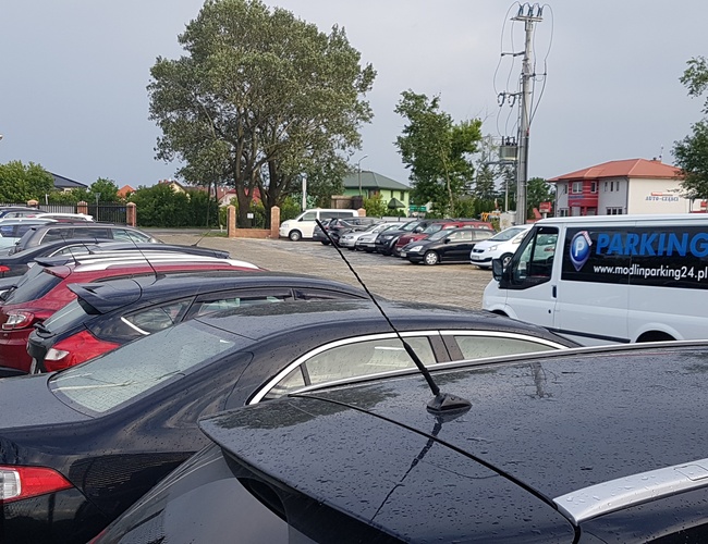 PARKING P24 PREMIUM - Na parkingu stoi wiele samochodów. Obok zaparkowano furgonetkę z reklamą. Teren jest otoczony drzewami i budynkami.