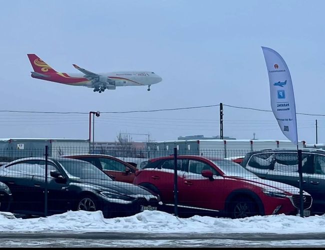 Parking Blisko Lotniska – Widoczna tablica reklamowa na trawie. W tle stoją samochody i reklamy. Pogodne niebo z chmurami.